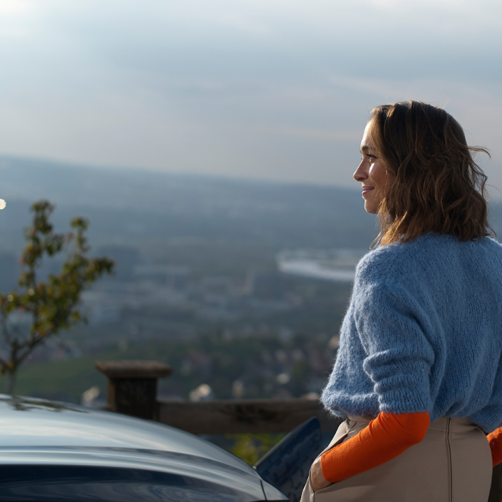 Mujer junto a un coche eléctrico oscuro, contemplando el paisaje desde un mirador al aire libre.